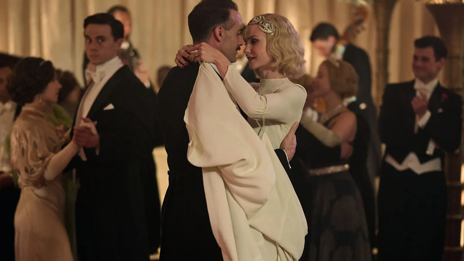 A blonde women in an impressive 1930s white dress dancing with a man with brown hair and mustache
