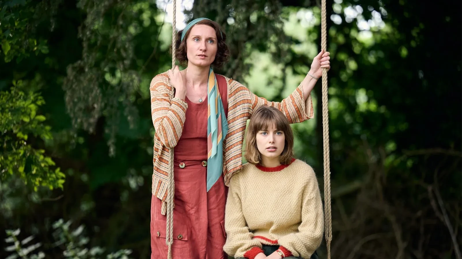 One young women in a pink 1930s style dress holding a rope swing where a younger women in a yellow jumper is sat