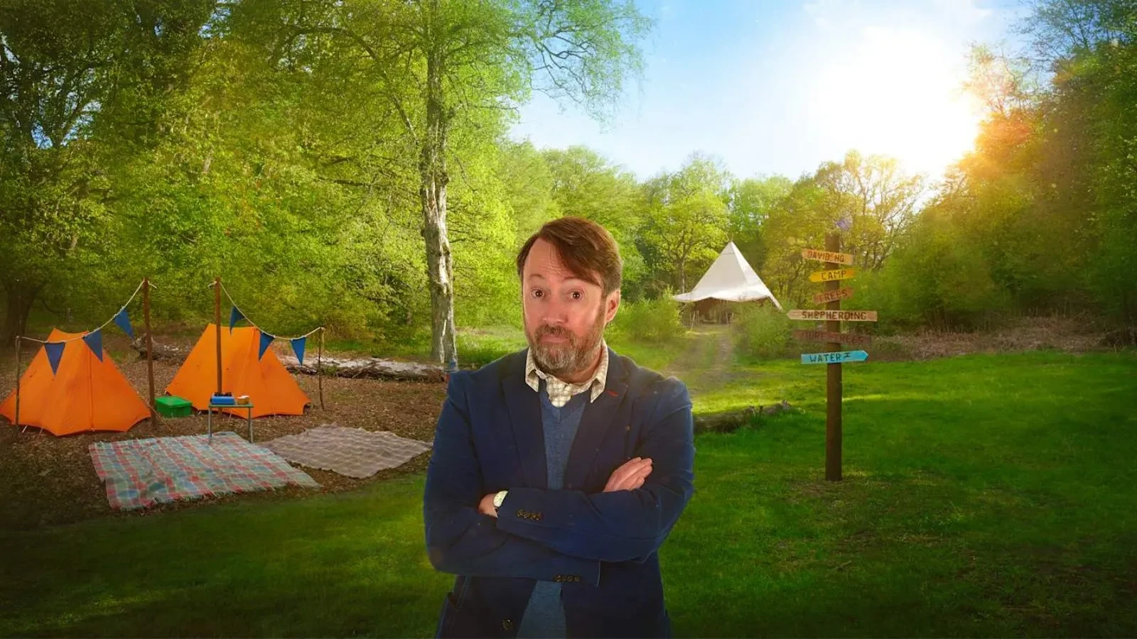 A man in a shirt with a smart jacket stood with tents and a forest in the background