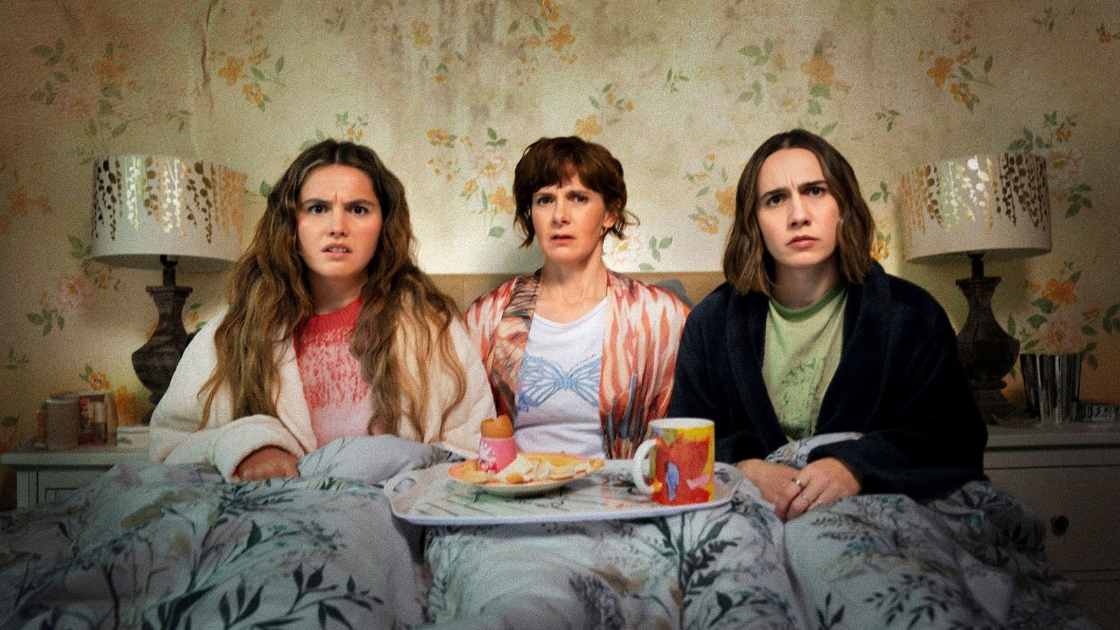 Two teenagers and their mum sat in a bed with a tray with egg and toast on