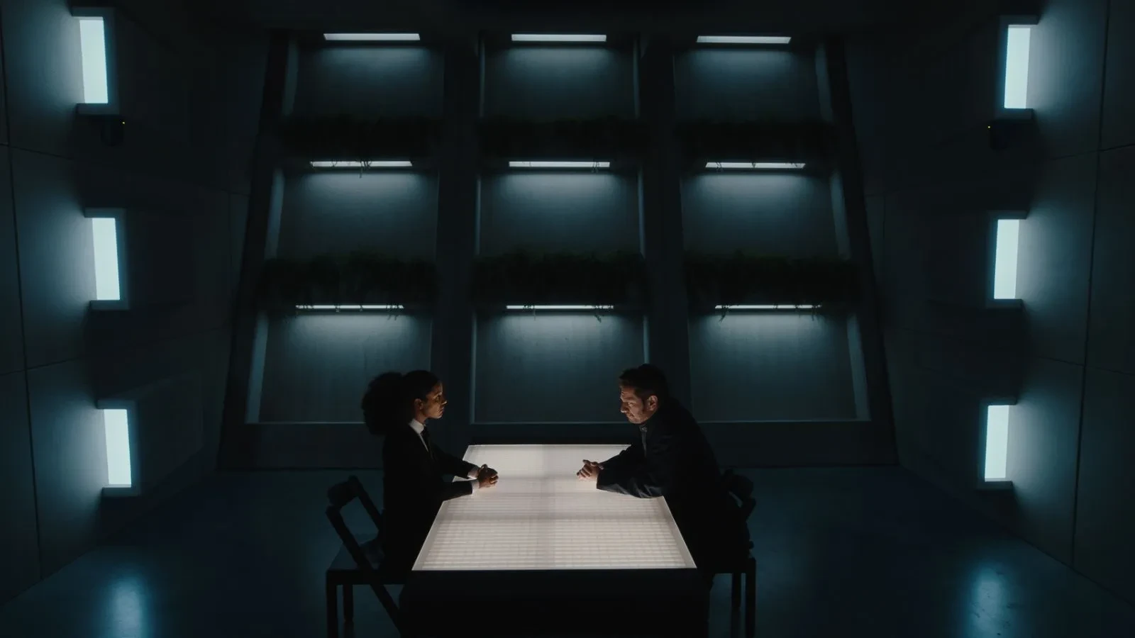 Two people in a dystopian looking room sat opposite each other at a table