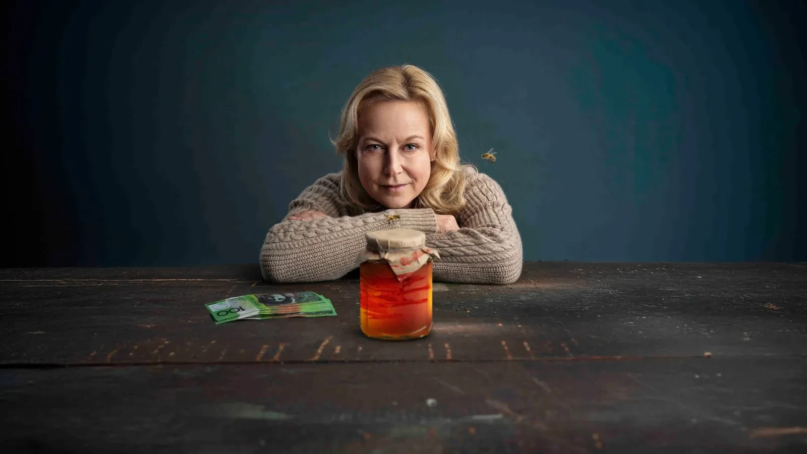 A blonde women wearing a woollen jumper with her arms on a table and a jar of something orange in front of her, smirking at the camera
