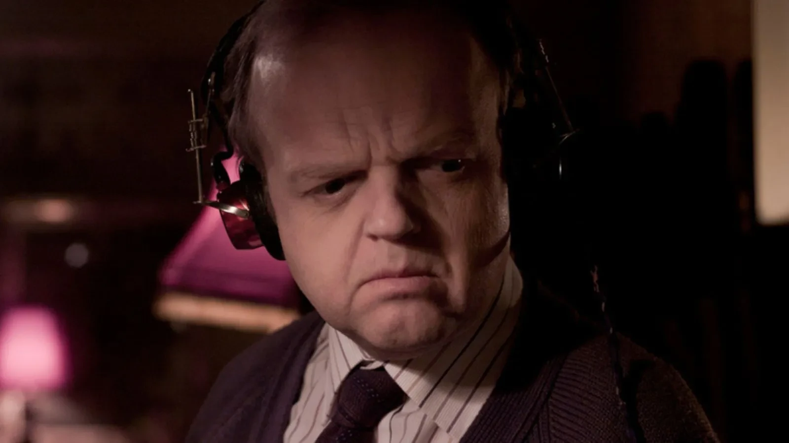 Close up of Toby Jones wearing a tie and a white striped shirt listening to something with black headphones on against a dark background