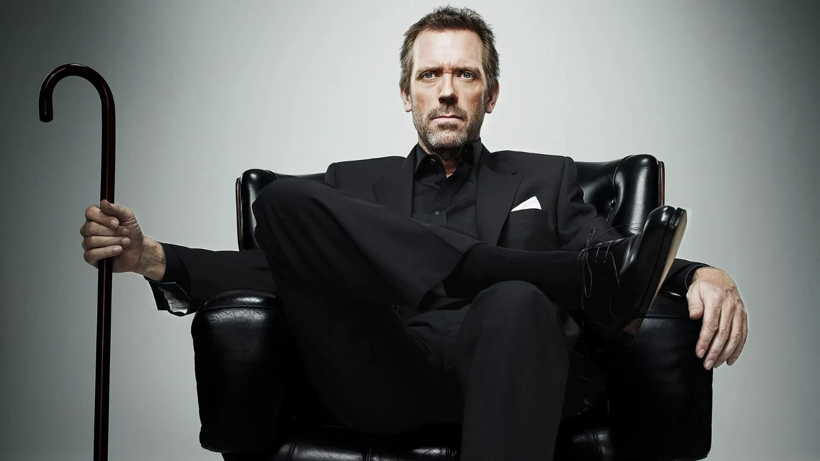 Hugh Laurie as Doctor House in the drama House sat on a chair wearing a black suit holding a walking stick