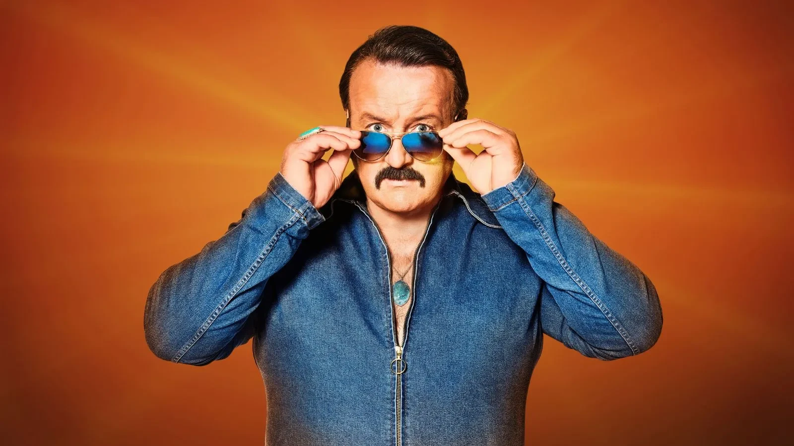 A man in the 80s wearing a denim outfit with slick hair and aviator sunglasses on stood against a plain orange background