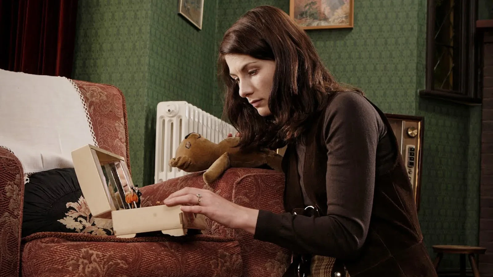 A Brunette women wearing a brown long sleeve top kneeling on the touching a jewellery box with her left hand and holding a teddy in her right