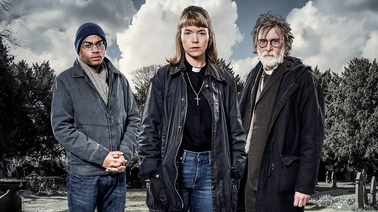 Anya Maxwell Martin wearing a clerical collar and a leather jacket stood with an older man with grey her to her right and younger man with glasses and a beanie to her left in a park.