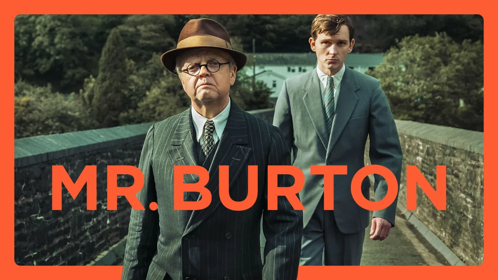 Toby Jones wearing a brown hat and black striped suit walking across a bridge with a younger man with brown hair in a grey suit walking behind him