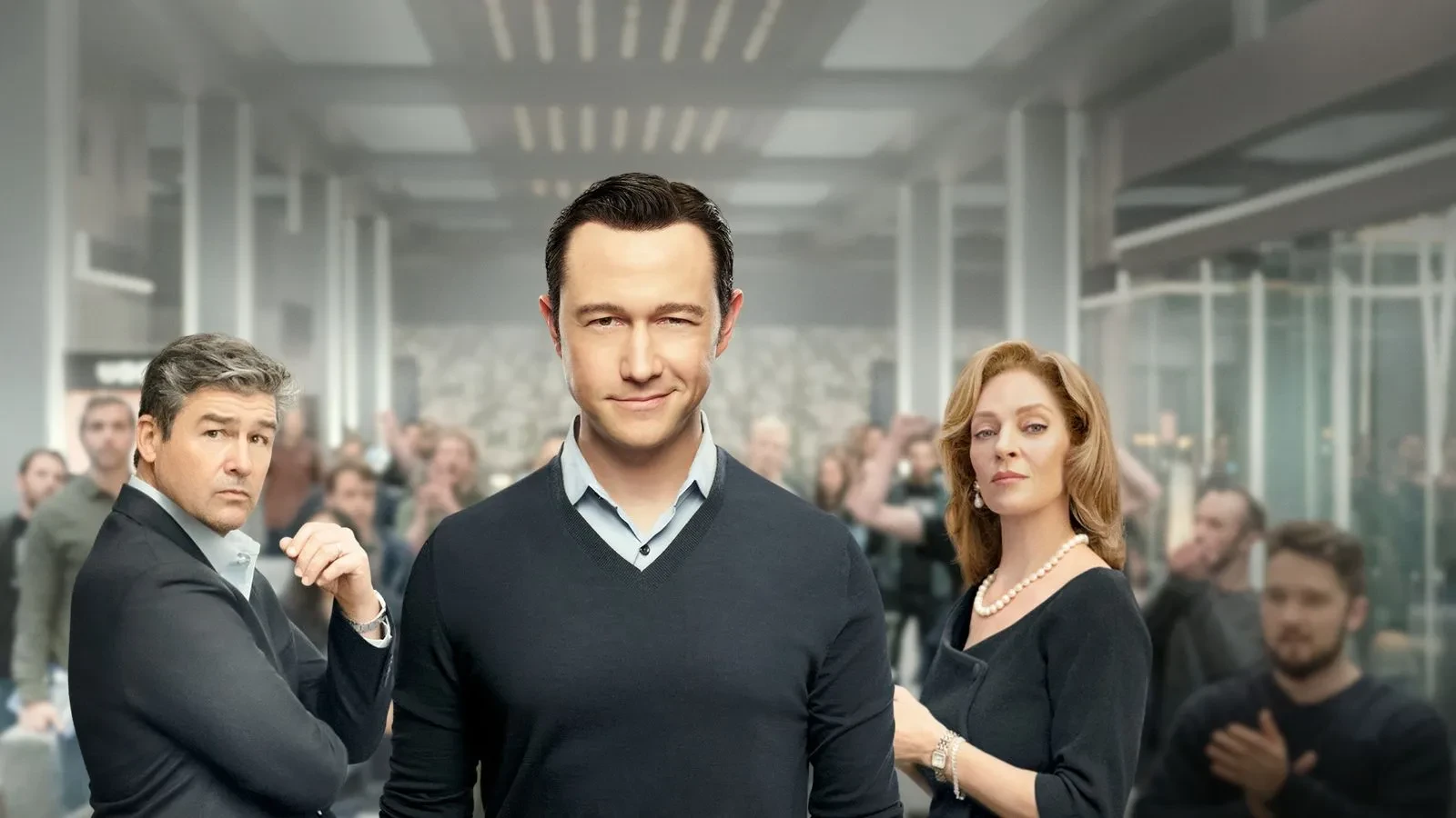 A man in a smart sweater and shirt smirking at the camera with a man and women also dressed smartly stood next to him and people cheering in the background