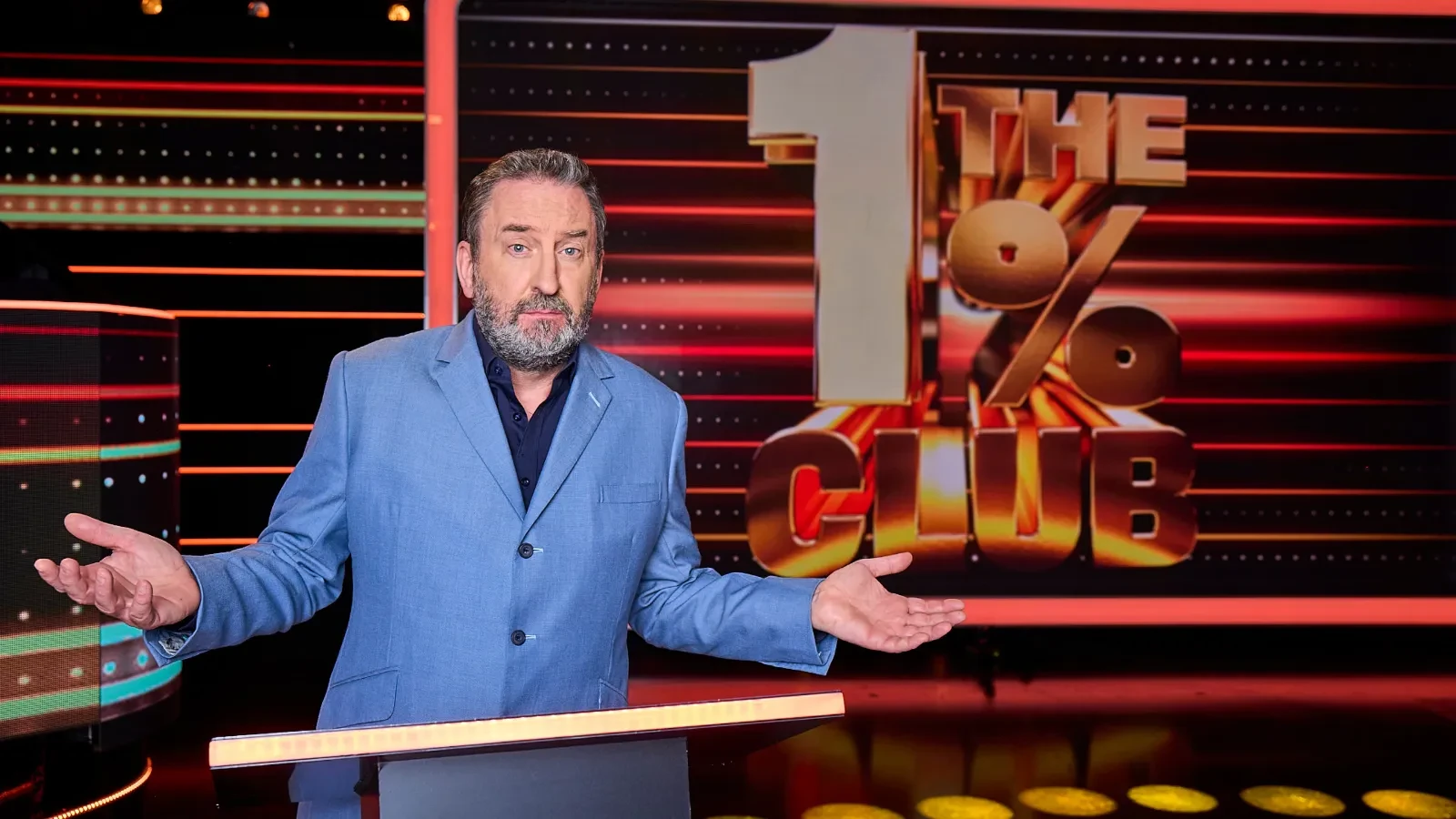 Person in a blue suit standing behind a podium on a brightly lit game show set with a large backdrop displaying ‘1 THE % CLUB’ in bold letters.
