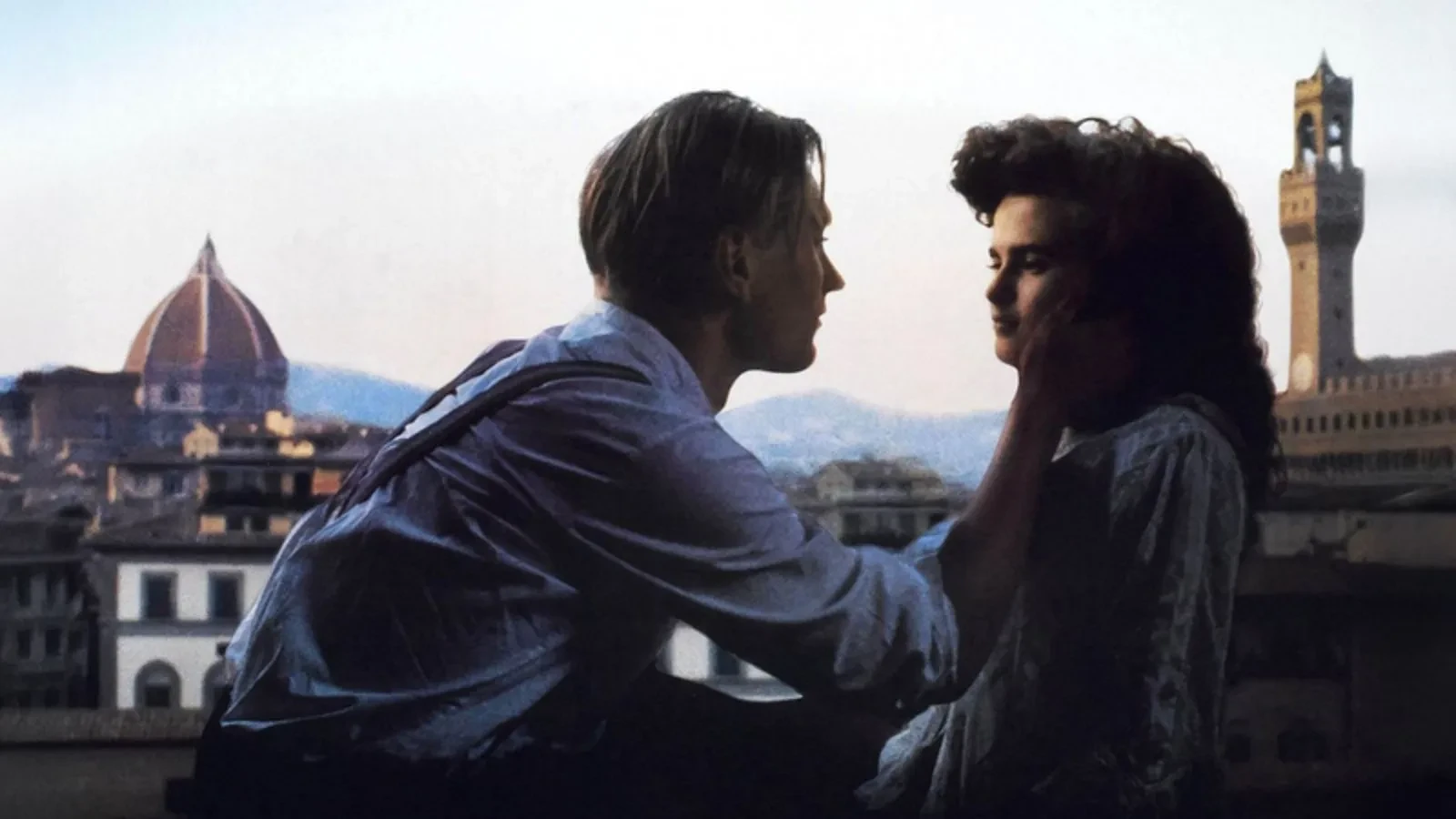 Still from the movie 'A Room with a View'. Two people sit close together on a balcony overlooking the historic cityscape of Florence, with the iconic dome of the Cathedral of Santa Maria del Fiore and a tall bell tower visible in the background, creating a romantic and scenic atmosphere.