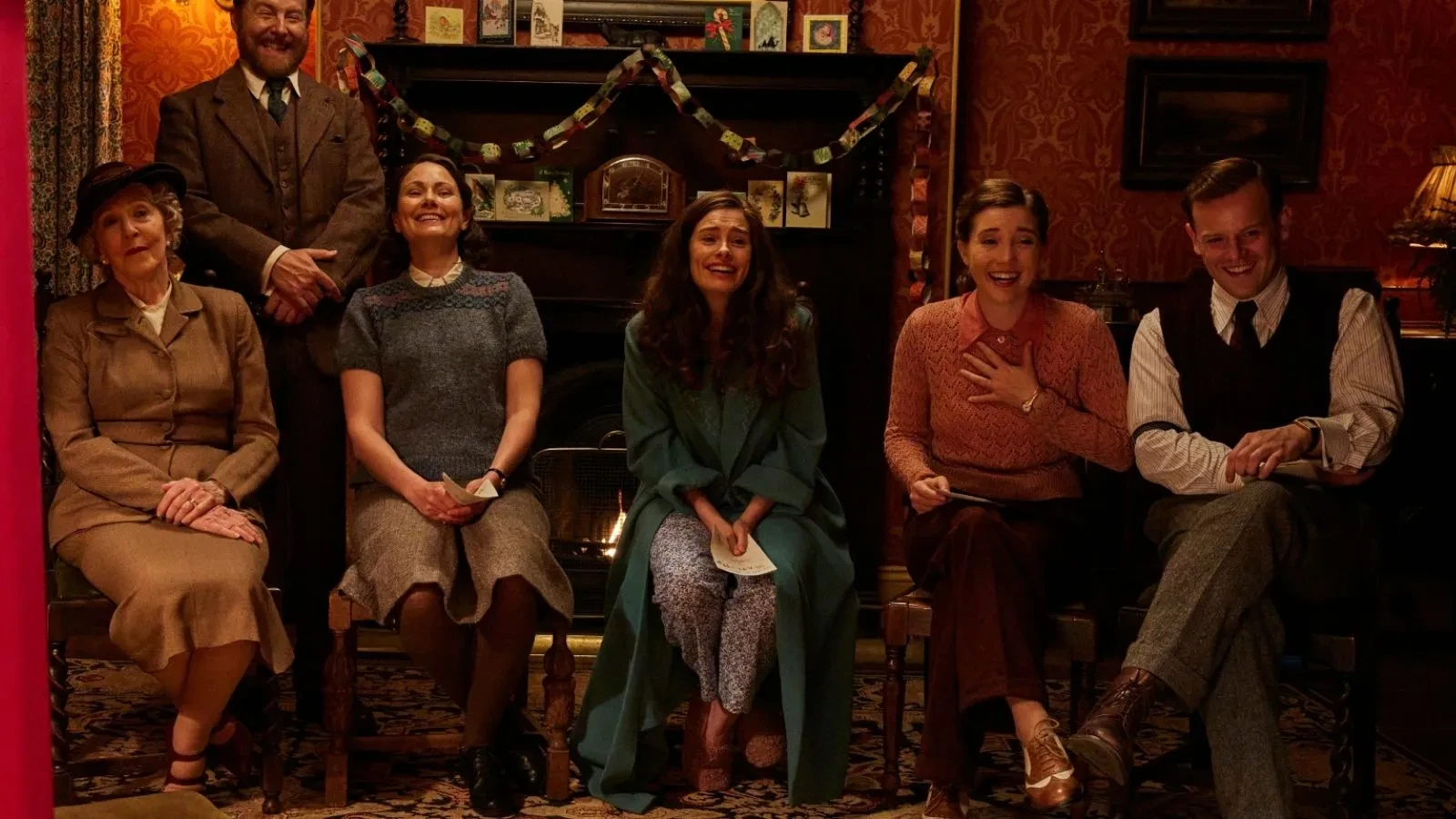 intage-style living room with a fireplace decorated with garlands, featuring people seated in period clothing on wooden chairs in a warmly lit setting.