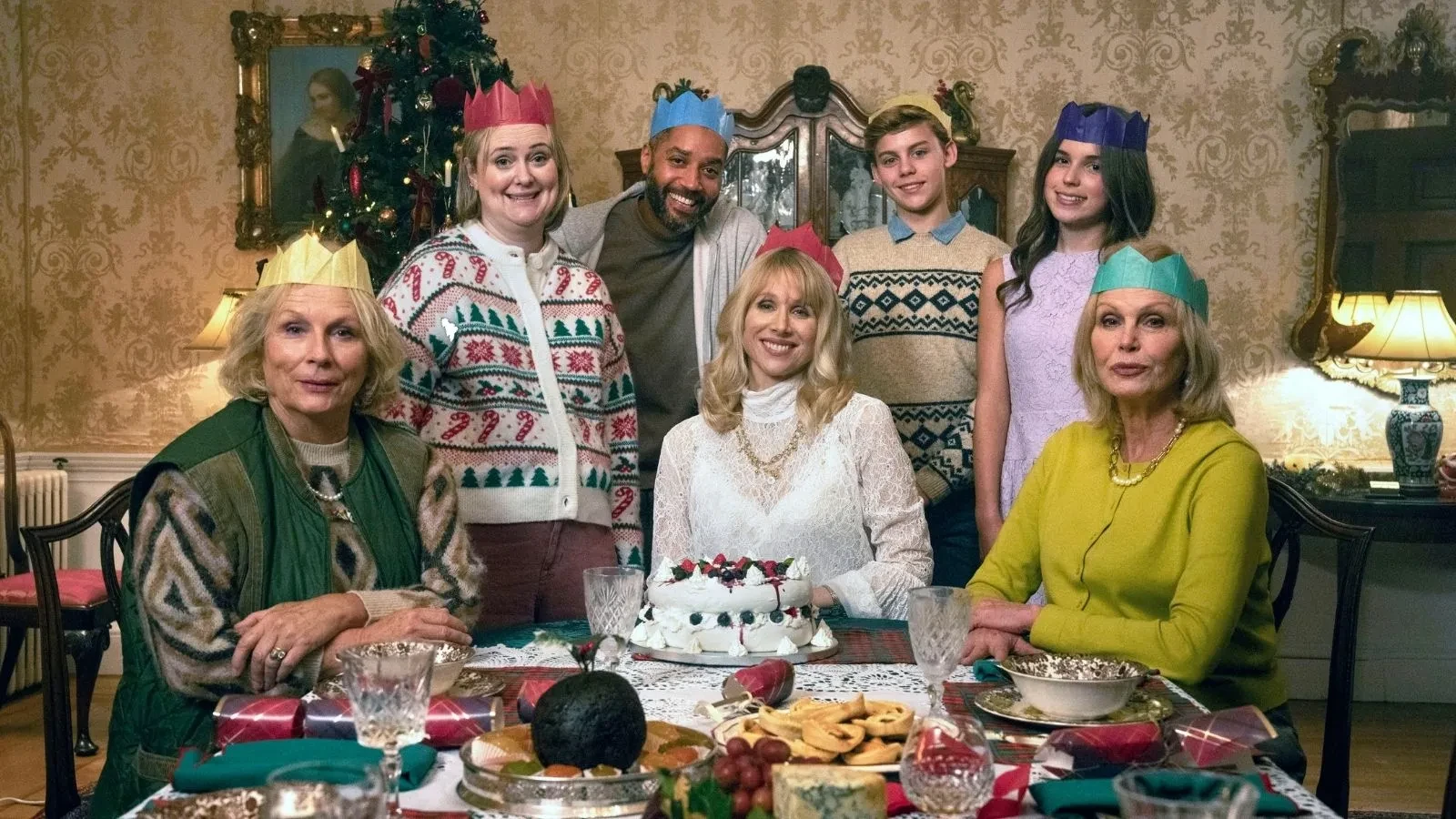 Festive dining room scene with a table set for Christmas, featuring a cake, crackers, and assorted treats, with people wearing colorful paper crowns and patterned sweaters.