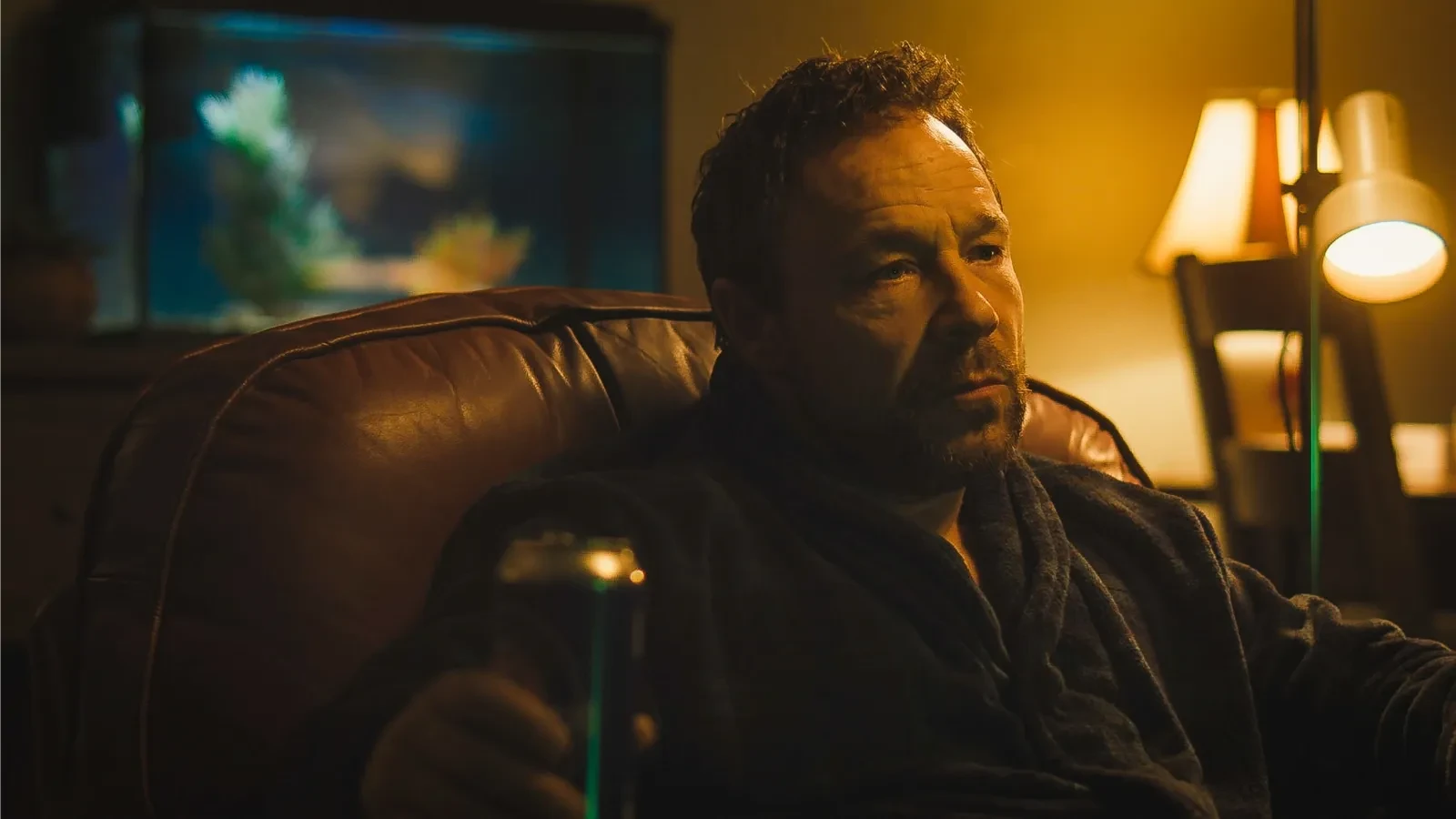 Still image for the 2021 film Boiling Point, showing a person sitting in a dimly lit room on a leather chair, holding a drink can. A fish tank is visible in the background, along with a lamp and part of a dining chair, creating a warm, moody atmosphere.