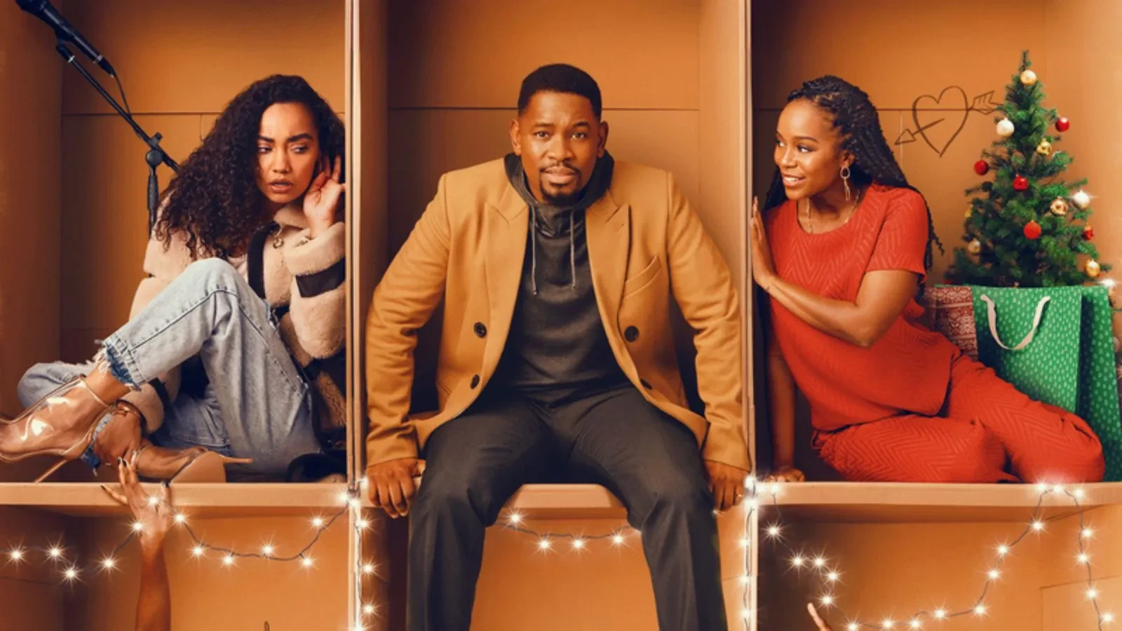 Three people sitting inside large cardboard boxes decorated with string lights; one box has a small Christmas tree and a green gift bag, while another has a microphone stand.”