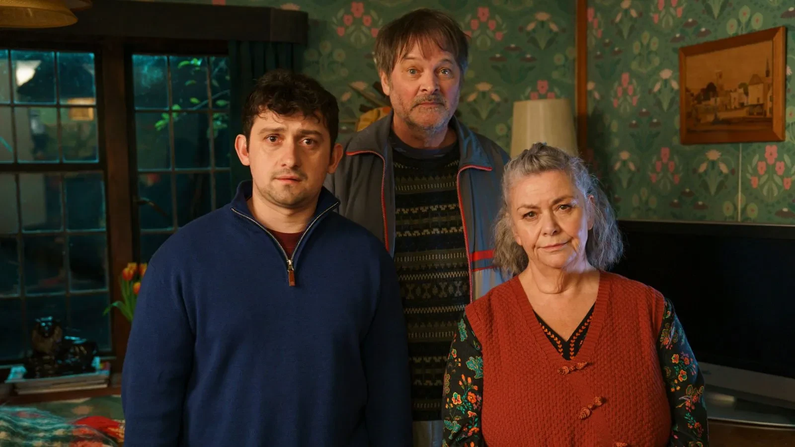 A still image from the British comedy drama series Can You Keep a Secret?, showing three people standing in a cosy living room with patterned green wallpaper, a large window, a television, and a lamp. One person wears a blue zip-up top, another a dark patterned jumper with a jacket, and the third a red knitted vest over a floral shirt.