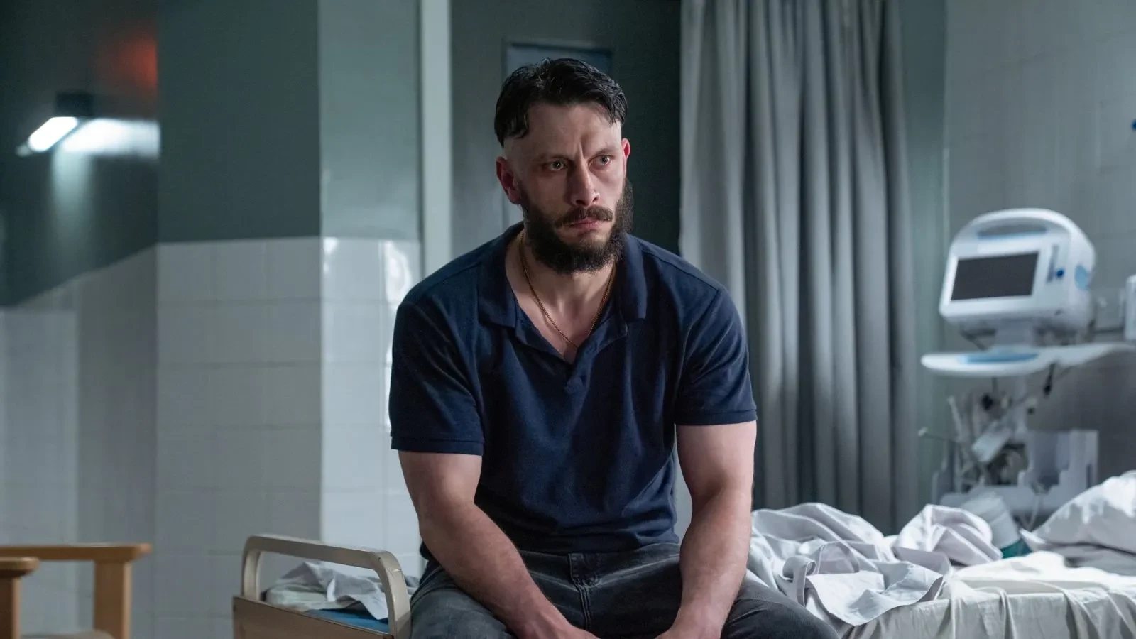 Hospital room scene from Richard Gadd’s drama “Half Man,” showing a person sitting on the edge of a bed with medical equipment and white sheets in the background.