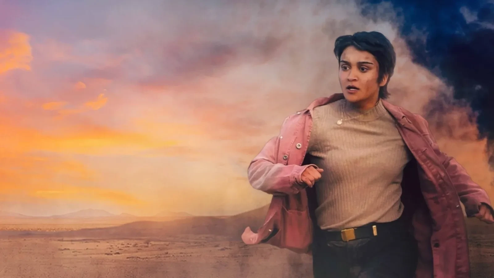 Promotional image for the 2022 film How to Blow Up a Pipeline, showing a person running across a dusty desert landscape at sunset, wearing a pink jacket and beige top. Dark smoke rises in the background against an orange and blue sky.