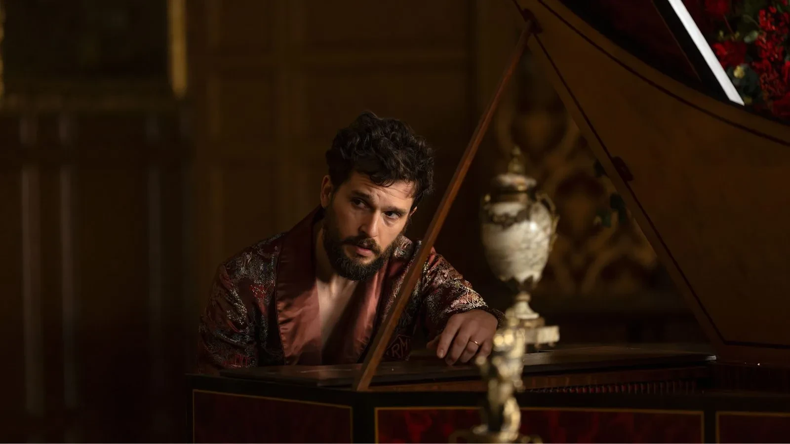 A still image from season 4 of the US drama series Industry, showing a person in an ornate patterned robe leaning on a grand piano in a richly decorated room with dark wood panelling and a decorative vase in the foreground.