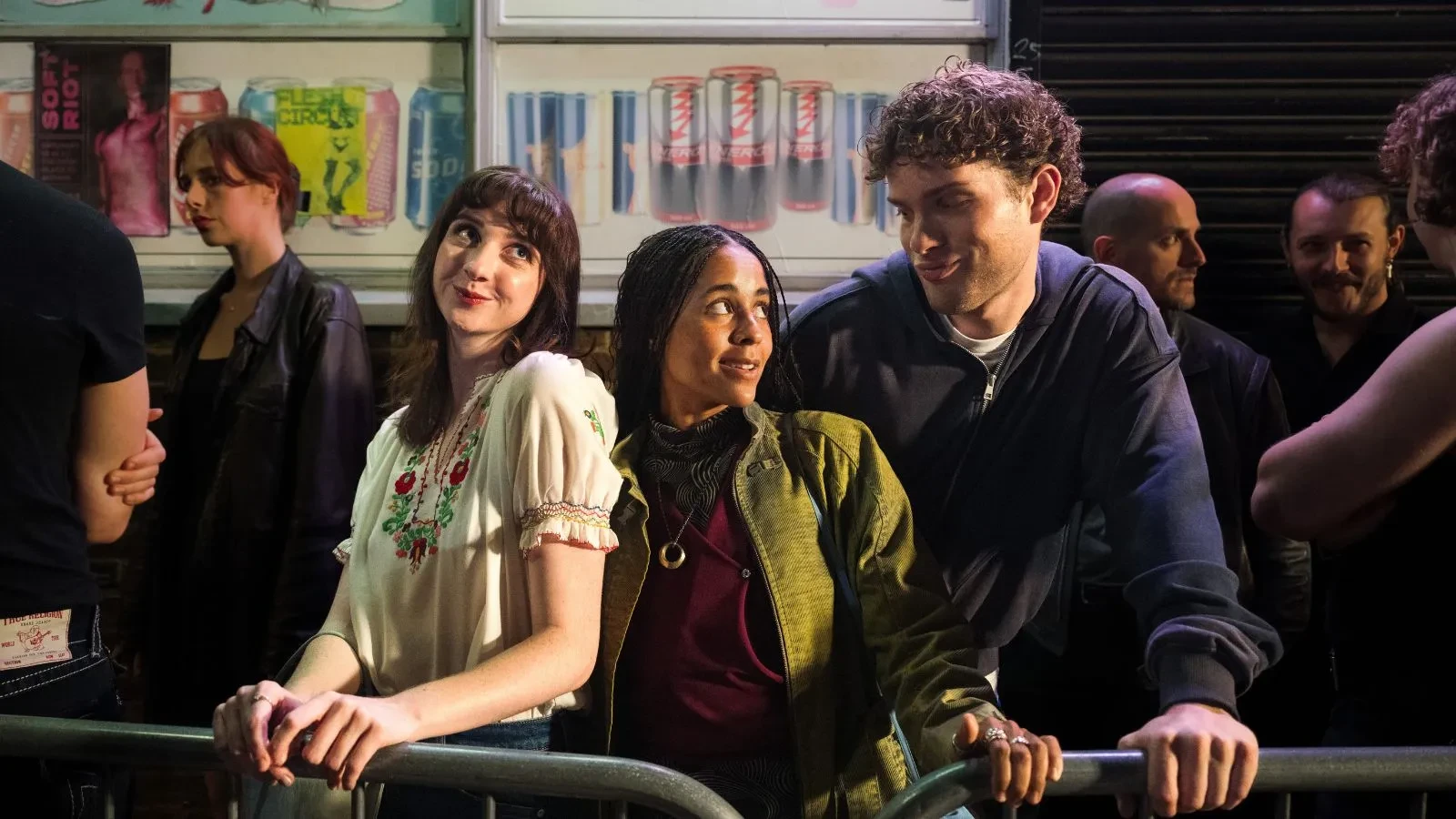 A nighttime street scene with several people standing behind metal barriers outside a venue. Bright lights illuminate the group, and a wall behind them displays posters and a row of drink cans.