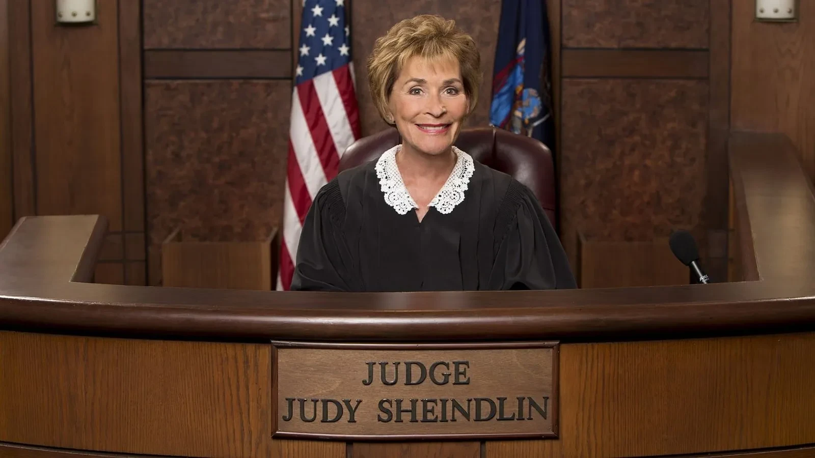 Promotional image from the courtroom reality show Judge Judy, showing a judge in a black robe with a white lace collar seated behind a wooden bench. The bench has a nameplate reading “Judge Judy Sheindlin,” and the background includes an American flag and a state flag.