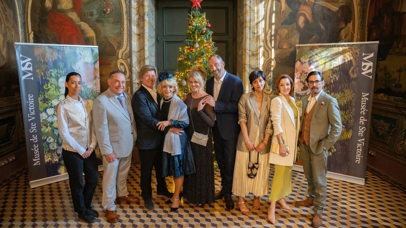 Group of people dressed in formal attire standing in an ornate room with patterned floor, a decorated Christmas tree, and banners reading ‘Musée de Ste Victoire.