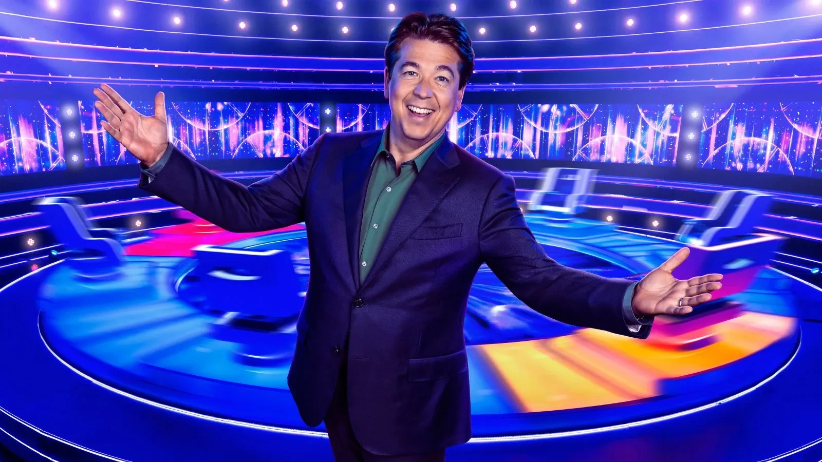 Game show set with bright neon lights and circular stage design, featuring a host in a dark suit standing with arms open in front of illuminated screens