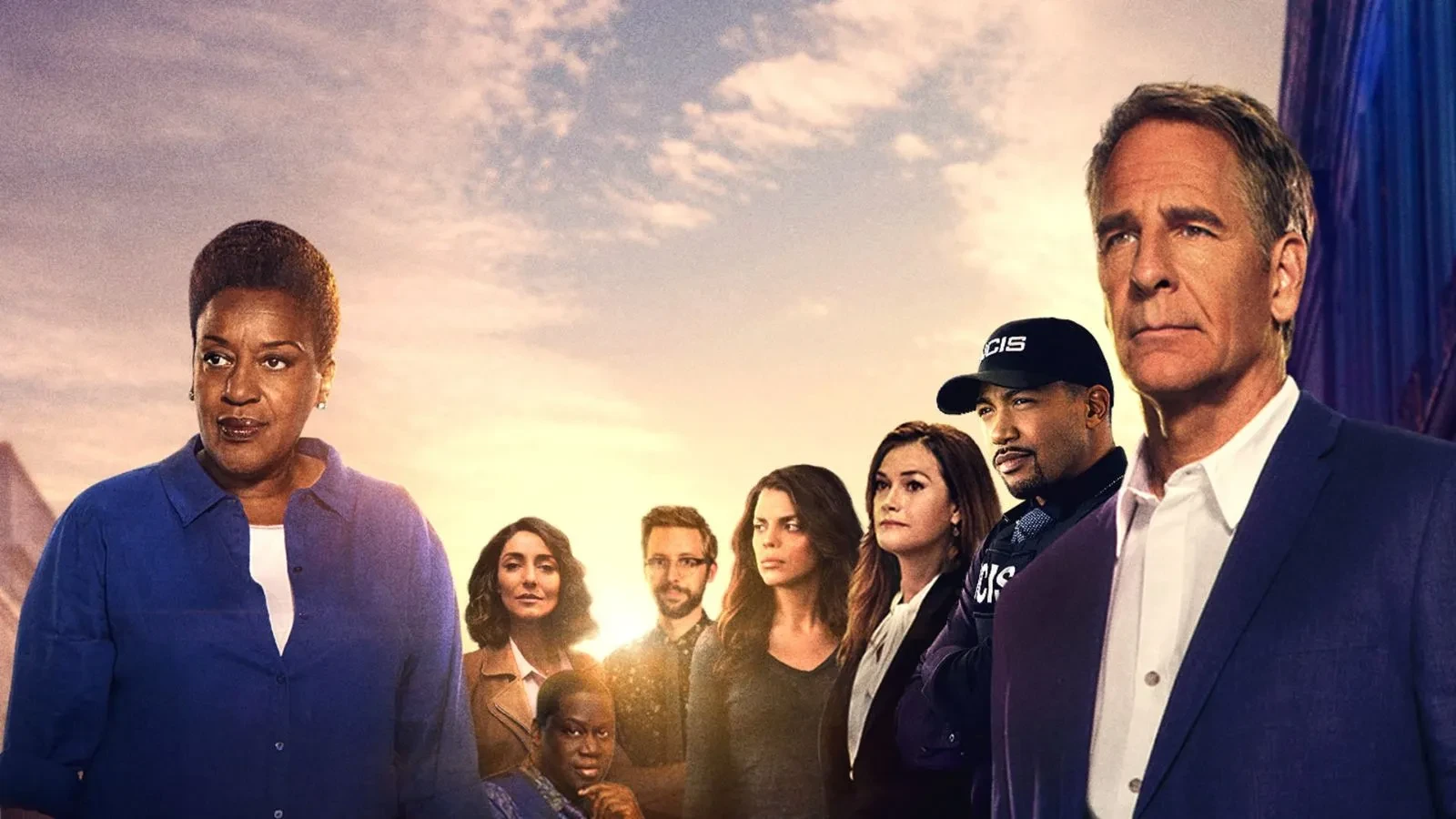 Promotional image from season 7 of the US crime drama series NCIS: New Orleans, showing a group of people standing together on a dock at sunset. The background features a calm sea, a boat, and a warm orange sky with scattered clouds. Some individuals are wearing casual clothing, while one has an NCIS cap and jacket.