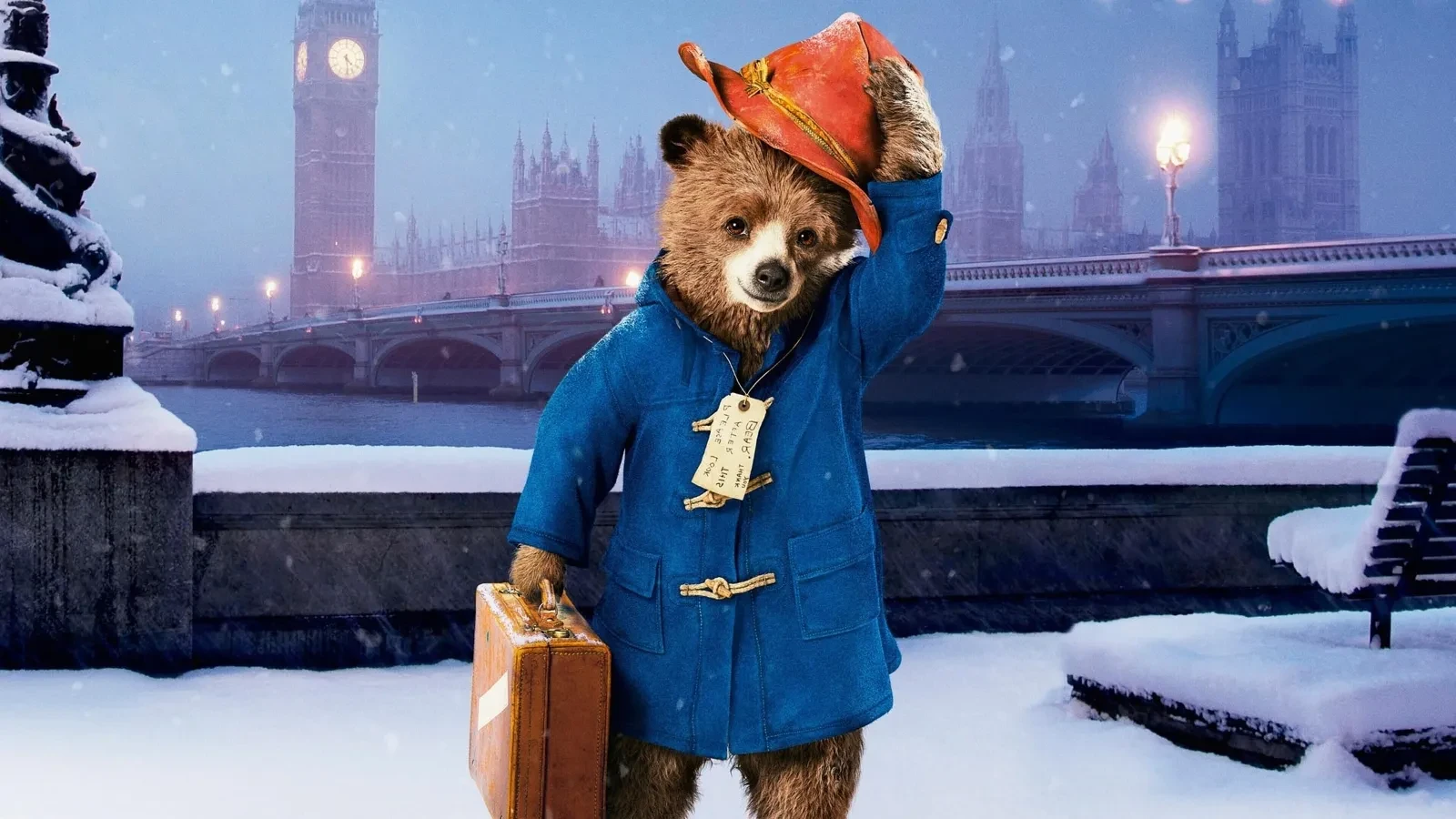 Promotional image for the 2014 film Paddington, showing the bear wearing a blue duffle coat and red hat, holding a suitcase in a snowy London setting with Big Ben and the Houses of Parliament in the background.
