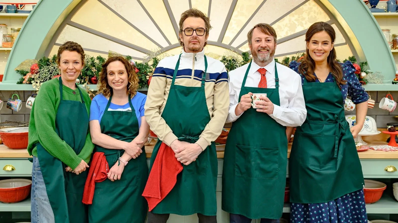 ive people wearing green aprons standing in a festive kitchen decorated with garlands and baking utensils.