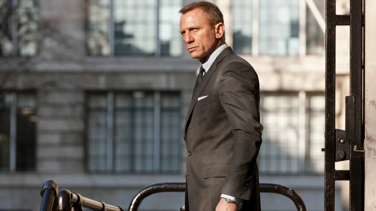 Still image from the 2012 film Skyfall, showing a person in a dark pinstripe suit standing outdoors near a metal railing, with a large building featuring tall windows in the background.