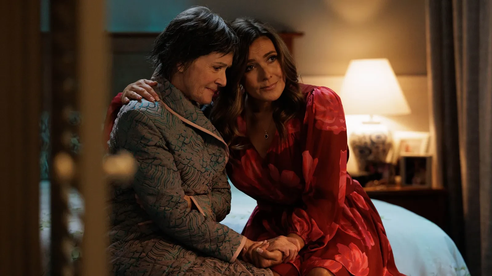 Two woman sat on a bed in a dimly lit room hugging.