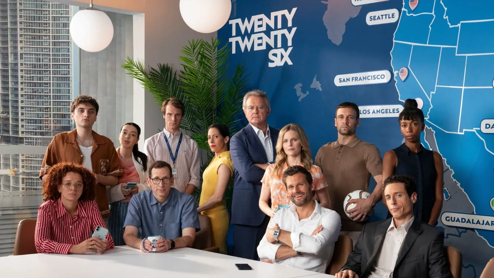 A group of people gathered around a modern conference table in a bright office. Behind them is a large blue wall map with city names like Seattle, San Francisco, Los Angeles, and Guadalajara, along with the text “TWENTY TWENTY SIX.” Two spherical pendant lights hang from the ceiling.