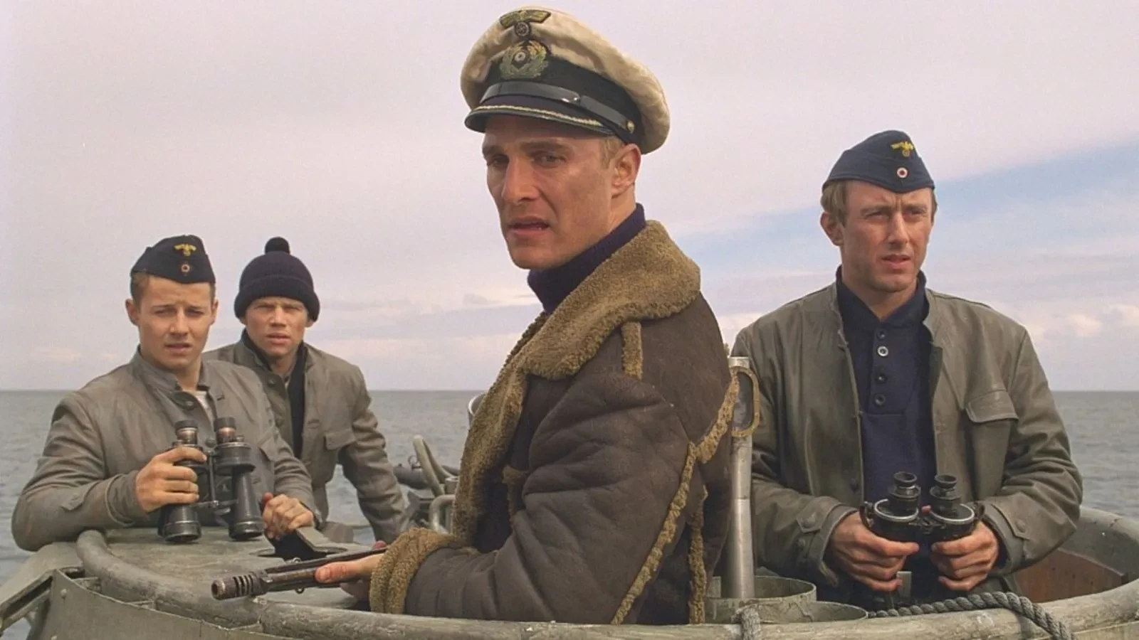 Still image for the 2000 film U-571, showing four naval crew members standing on the deck of a submarine at sea. They are dressed in military uniforms and caps, with some holding binoculars. The ocean and cloudy sky form the background.
