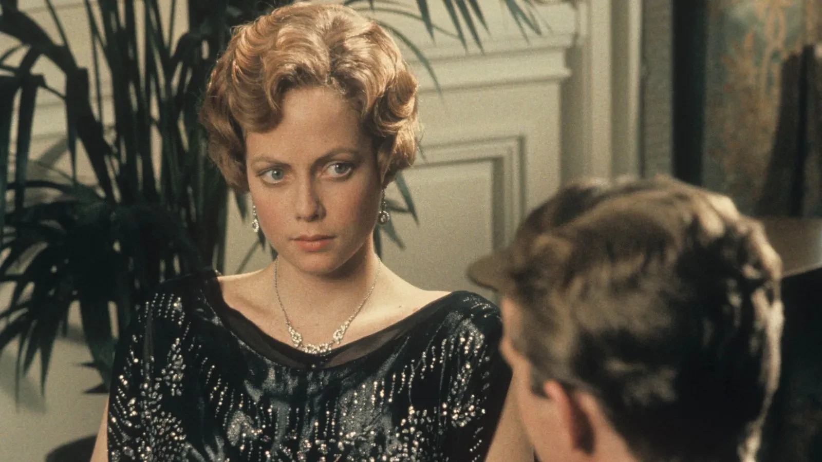 Person wearing a black sequinned dress with a necklace, seated indoors with plants and panelled walls in the background.