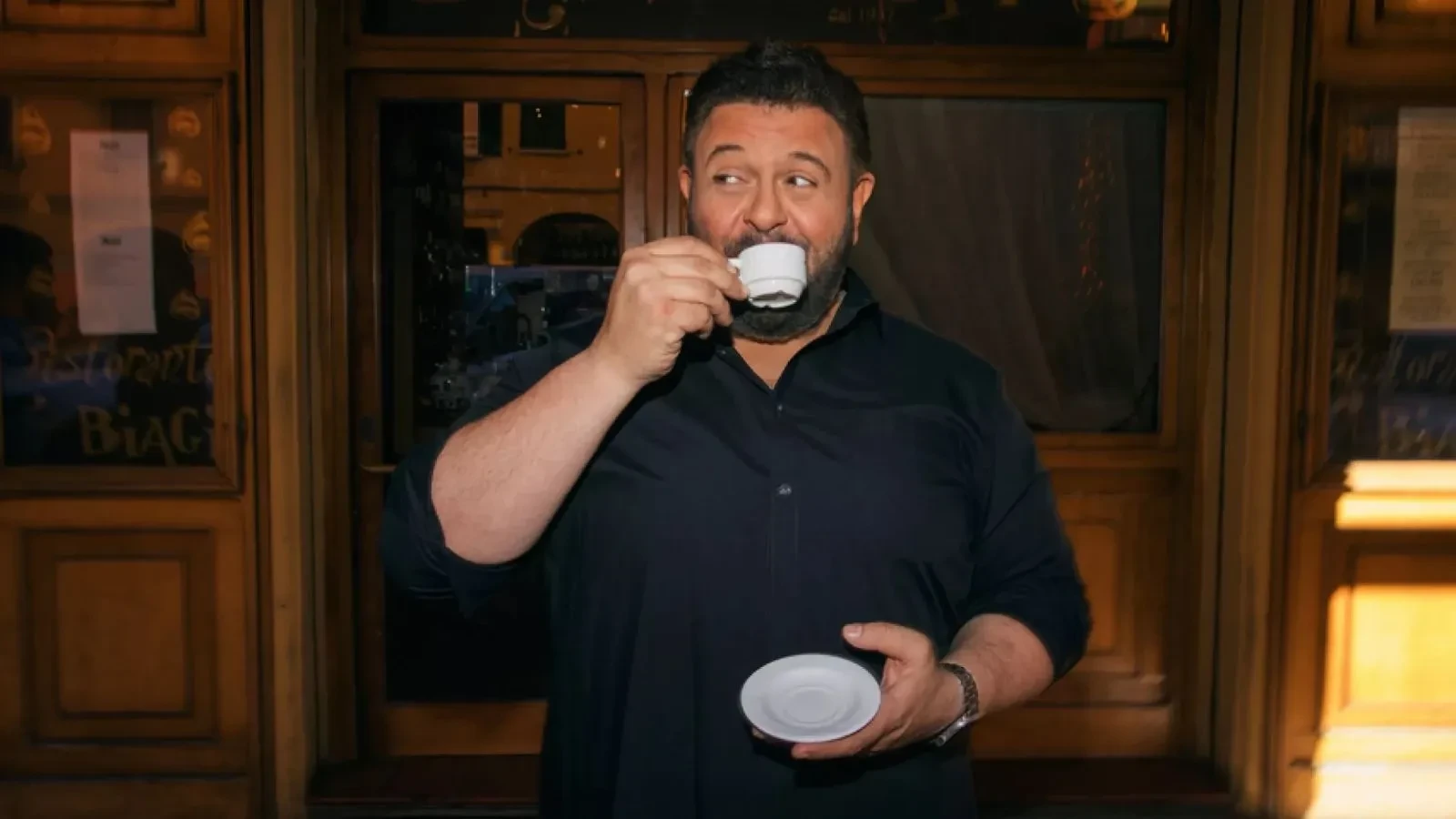 A person wearing a dark shirt is standing outside a wooden doorway, holding a small white saucer in one hand and a tiny cup in the other, as if drinking espresso.
