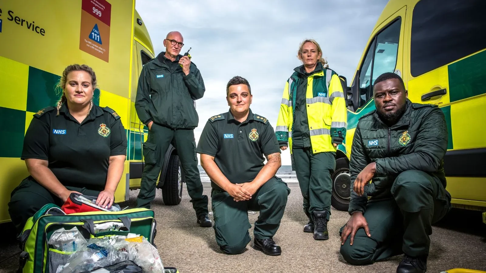 Five paramedics in green NHS uniforms are positioned between two yellow and green ambulances. Medical equipment bags are open on the ground in front of them, and one person is holding a radio. The scene appears to be outdoors on a road or pavement.