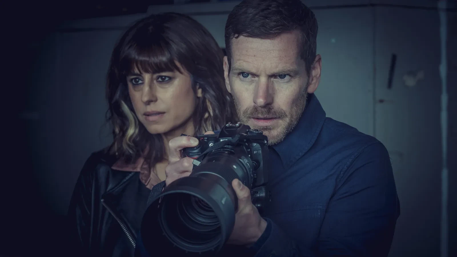 Two people holding a professional camera with a large telephoto lens in a low‑light environment.