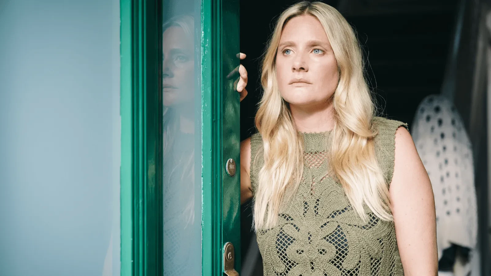 Woman with long blonde hair standing in a doorway, wearing a green patterned sleeveless top.