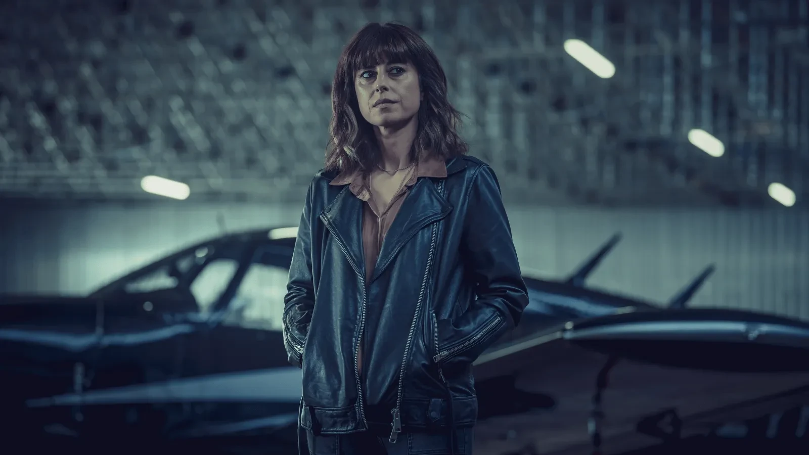 Woman wearing a black leather jacket standing in an aircraft hangar beside a small plane.