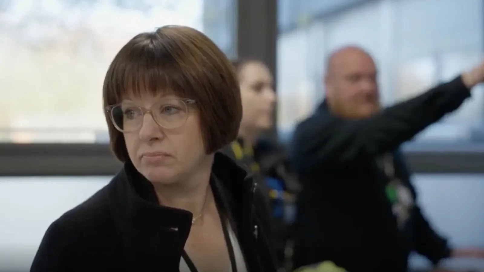 A person in a dark coat stands indoors near large windows while another person in the background gestures toward something out of frame.