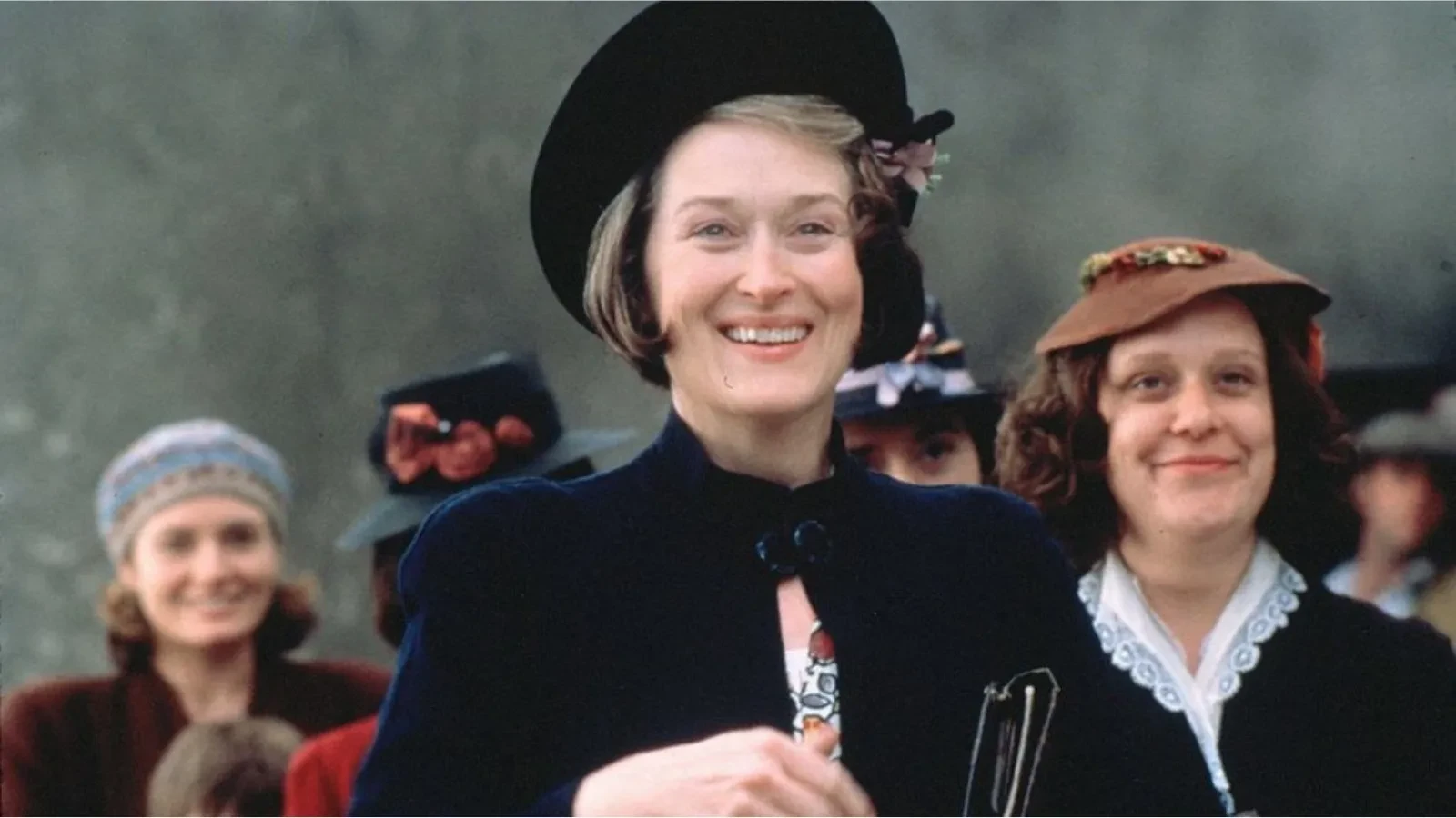 A group of women dressed in 1930s or 1940s period clothing, wearing vintage hats adorned with flowers and ribbons, standing together in an outdoor setting with a muted, textured background.