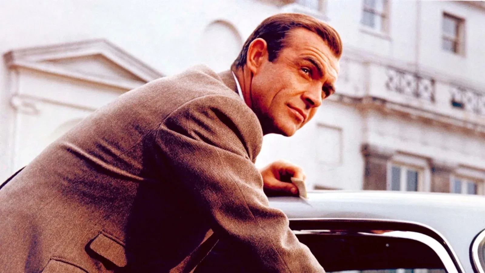 A man in a brown suit leans over the roof of a classic car, set against the backdrop of an elegant white building with traditional architectural details.