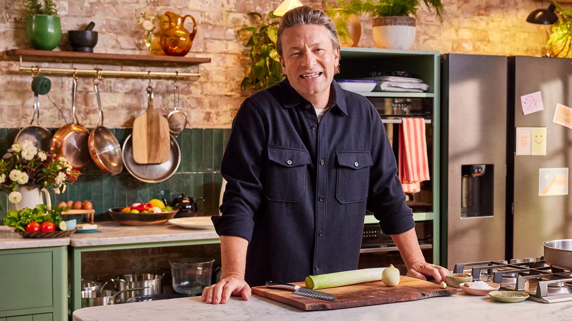A person standing in a rustic kitchen, leaning on a marble worktop beside a chopping board with a leek, onion and knife, surrounded by fresh vegetables, copper pans and modern cooking equipment.