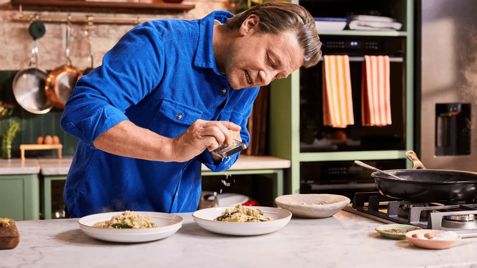 A person in a bright blue shirt grates an ingredient over two plated dishes in a kitchen, with pots, bowls, and cooking utensils around them.