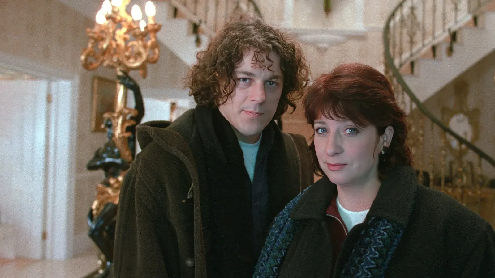 Two people standing inside an elegant, ornately decorated interior featuring a grand staircase, golden detailing and a large decorative lamp, creating a sophisticated and slightly mysterious atmosphere.