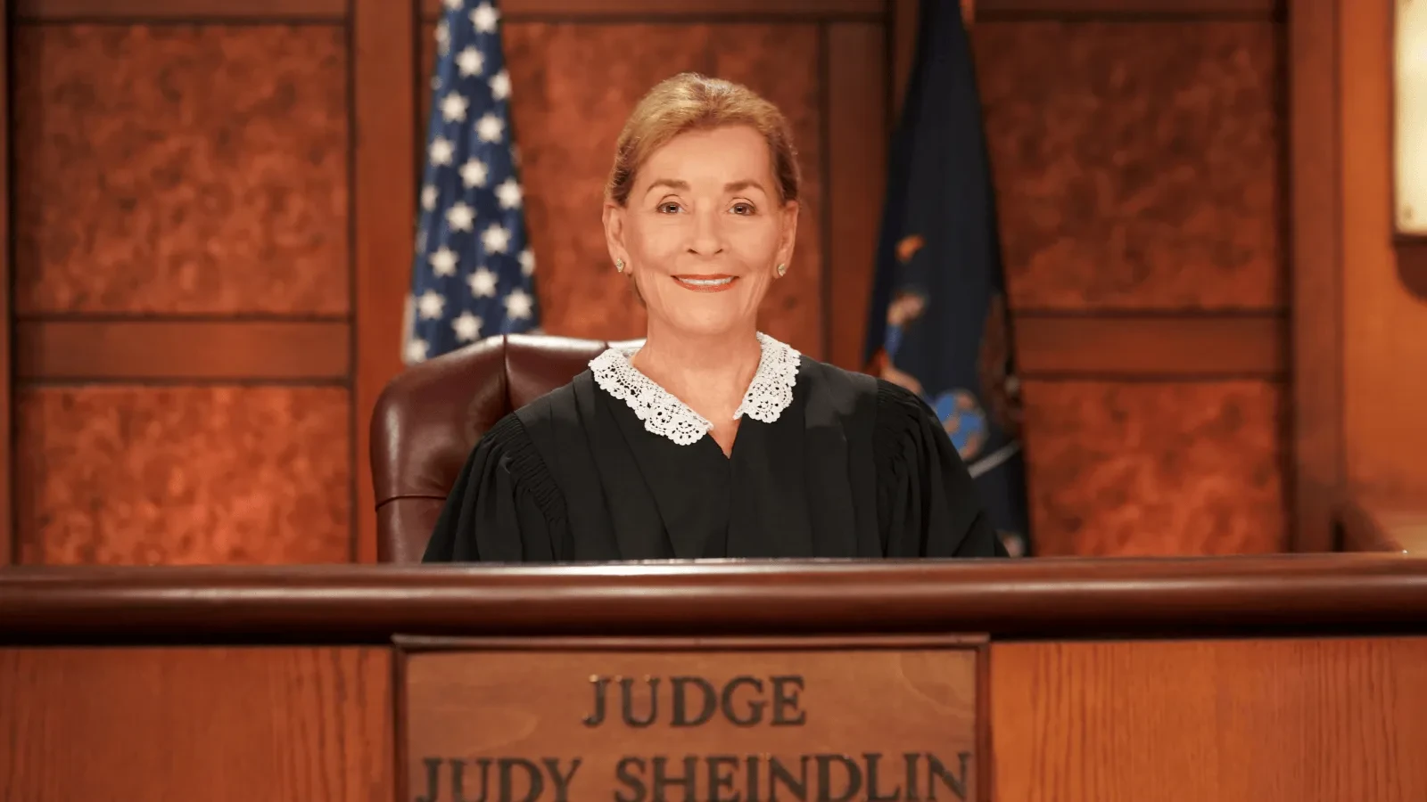A judge wearing a black robe with a white lace collar sits at a courtroom bench. Behind the judge are wooden panels and two flags. The front of the bench displays a nameplate reading ‘Judge Judy Sheindlin.