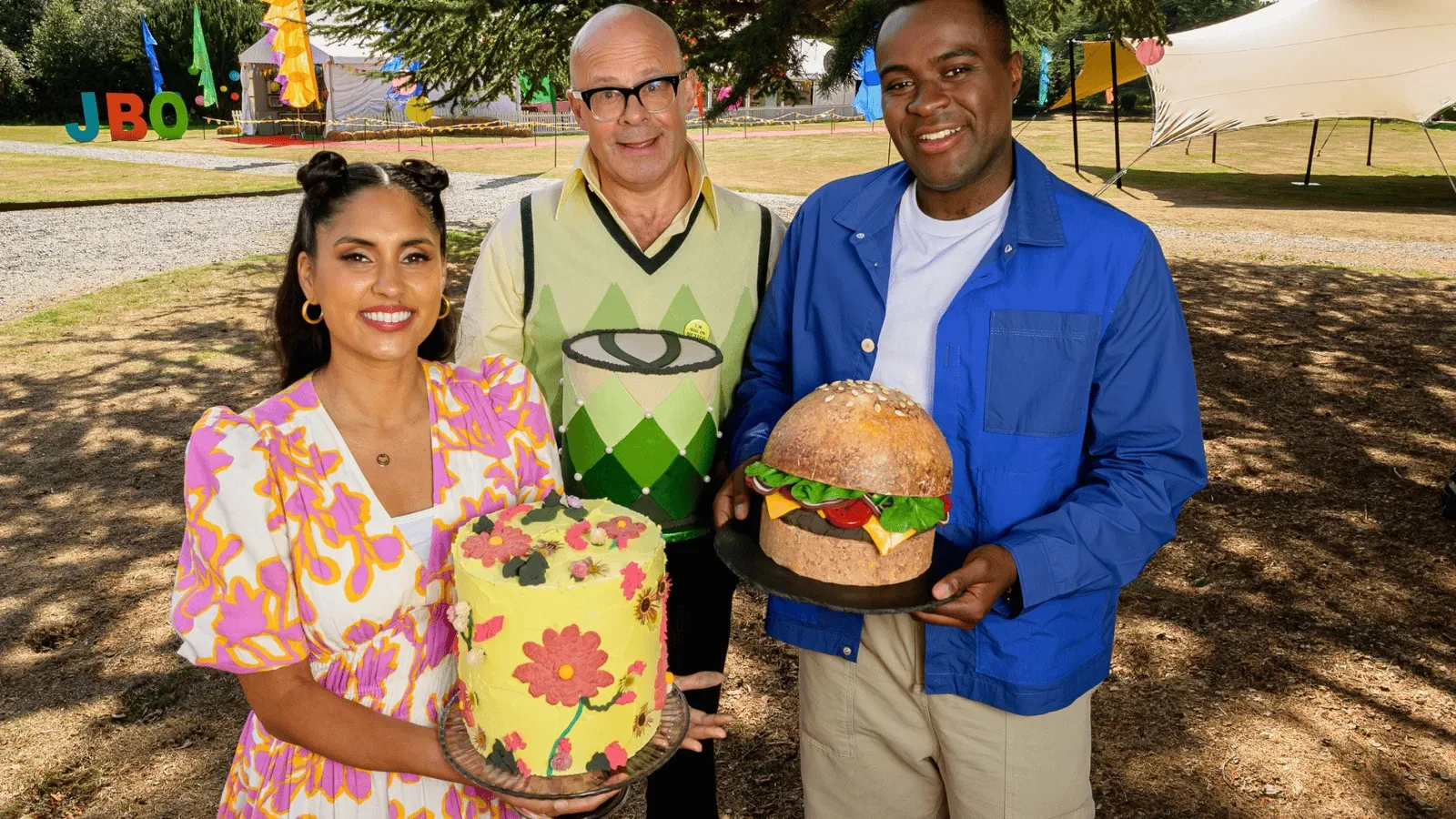Three individuals outdoors hold colourful, creatively decorated cakes, with bright festival-style decorations and a large tent visible in the background.