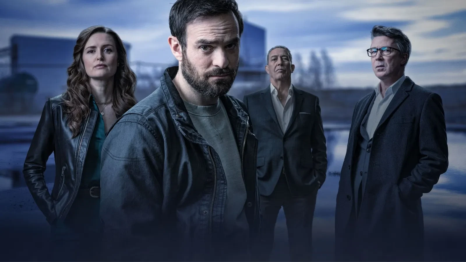 A group of four people standing together in a moody, industrial outdoor setting at dusk, dressed in dark, stylish clothing including leather jackets, casual layers and tailored suits, creating a tense and atmospheric crime‑drama aesthetic.