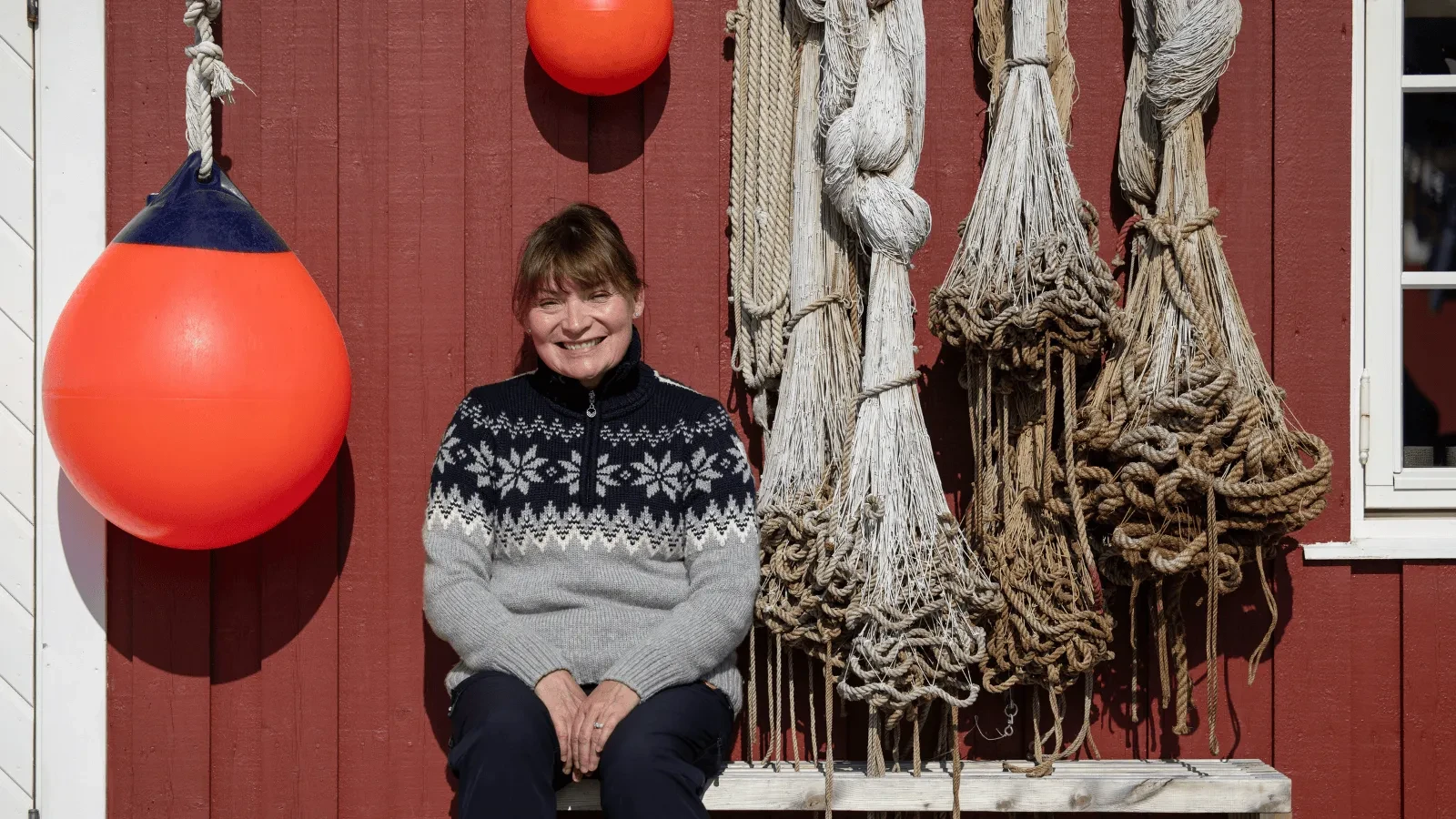 A person wearing a patterned knit sweater sits on a bench against a red wooden wall, surrounded by fishing buoys and bundles of coiled rope.